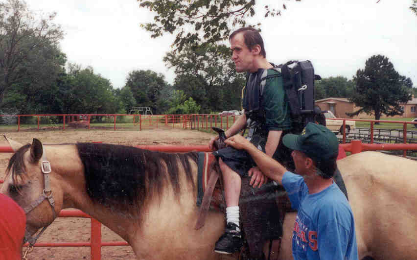 A Client riding a horse using the adaptive saddle device assisted by a staff member.